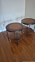 Front view of two wooden circular side tables with lower shelves on a hardwood floor.