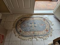 Photo of a small oval blue rug with a floral pattern and fringe placed on a tiled floor near a door.