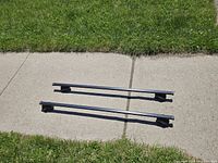 Two assembled roof rack crossbars viewed from above on pavement and grass background.