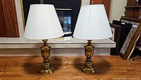 Two brass table lamps together on a surface under a mantle, in front of a fireplace. Both with pleated white lampshades.