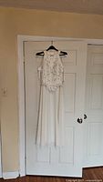 White sleeveless medium size dress with a sequined top overlay hanging on a door. Skirt is plain white and flowing.