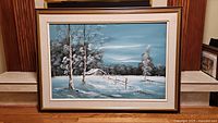 A framed oil painting on canvas showing a winter farmhouse scene with snow, trees, fence, and a farmhouse in the background.