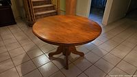 Photo showing a round wood kitchen table with a warm medium-brown finish, placed on a tiled floor next to stairs and hallway.