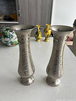 Pair of silver metal vases with engraved floral designs, shown side by side on white surface with other items blurred in background