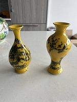 Two yellow ceramic vases with black floral designs side by side on a white surface.