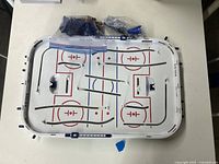 Unassembled white rectangular hockey game surface with rink markings and metal rods attached.