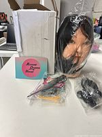 Mannequin head in plastic wrap with brand card and packaging box visible, along with some styling accessories in bag.