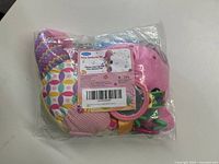 Soft baby plush toy packaged in transparent plastic bag, showing various colorful fabric patches and attached teething rings from different angles.