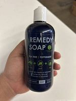 Front view of 12 oz dark blue bottle labeled Remedy Soap with listed ingredients and intended use for foot and body. Identifies key essentials: tea tree, peppermint, aloe.