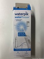 Box front showing Waterpik Cordless Express Water Flosser branding, product image, features like cordless convenience, ADA accepted, and inclusion of 2 tips.