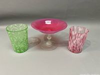 Front view of three colored art glass items: one green tumbler with white pattern, one pink tumbler with white twisted pattern, and one pink comport with clear stem and base