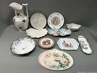 Set of various porcelain items arranged showing pitcher, trays, plates, and gravy boat with floral and scenic designs.