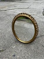 Full view of oval ornate wall mirror standing on cracked concrete surface, showing decorative gold frame and clear reflective glass.