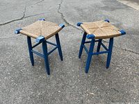 Pair of two blue painted wooden stools with woven rush seats shown standing on pavement.