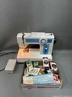 White Deluxe Sewing Machine Model 1510 with multiple sewing accessories in a box below it. Machine has power cord and foot pedal attached.