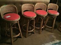 Four rattan swivel bar stools with red vinyl round seats arranged in a row against a sofa, showing overall items and condition including a ripped seat.
