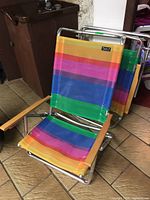 Two fold-up metal frame beach chairs with colorful striped canvas seating and wooden armrests stacked and one chair open on tiled floor.