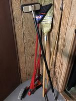 Photo showing three cleaning tools leaning against a wooden paneled wall including a floor brush, black bristle broom, and traditional straw broom.