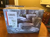 Boxed Canvas Wild Rose porcelain dinnerware set with floral pattern featuring stacked plates, bowls, and mugs shown on packaging.