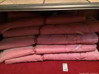 Stack of six reddish patio chair cushions placed on red carpet with visible tie straps and weathered condition.