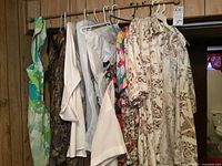 Photo showing various vintage clothes hanging on hangers including floral apron, white blouse, camouflage pattern garment, floral skirts and dresses with abstract and geometric prints.