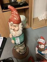 Full view of vintage gnome sculpture standing on a circular brown base, showing weathered red hat and green jacket.
