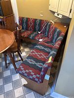 Wooden corner banquette with upholstered seat and back in geometric red, blue, and black fabric, shown in kitchen corner next to wooden table and chairs.