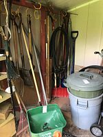 Photo showing garden tools including rakes, hoes, shovels, hoses arranged upright in a shed, plus the green Scotts broadcast spreader on floor
