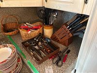 Photo of kitchen countertop showing Henckel knife block with knives, stoneware utensil holder filled with metal kitchen utensils including a whisk and spoons, wicker baskets holding additional utensils and a tray of stainless steel flatware including spoons and forks