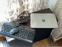 HP Pavilion laptop closed on table, Logitech keyboard and mouse visible with power adapter cords. Surrounding includes furniture and lace curtains.