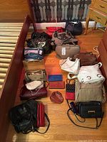 Overview photo showing 15 handbags and wallets in various colors and materials arranged on a hardwood floor.