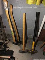 Four wooden-handled hand tools standing against a gray wall: two axes with natural wood handles (one curved, one straight) and two mallets, one with a black and yellow head, one with a plain wooden head.