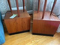 Pair of mid-century modern teak stands with black leather bases shown on a wood floor in front of lace curtains