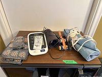 Full view of all items on a wooden surface including Omron blood pressure monitor, cuff, wooden massager, blue heating pad, and hearing aid batteries.