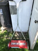 Front view of the Noma Easyclip push lawnmower on grass against a white wall, showing the red cutting unit and metal handle with black grips.