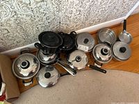 Photo of a set of assorted pots and pans including stainless steel and black cookware with lids arranged on a carpeted floor near a wall.