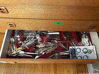 Drawer view showing assorted flatware including forks, spoons, knives, and cooking utensils with a card of beverage charms included.