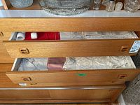 Two open wooden drawers containing napkin rings, folded red and white napkins, and several folded tablecloths with lace and embroidery detail.