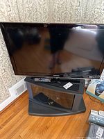 Samsung 40-inch TV displayed on the black TV stand with glass door, remote control placed on stand shelf, wooden floor background.