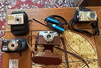 Lot overview with all items: SVS camera in leather case, Sony Cyber-shot, Nikon Coolpix, Suntone radio headset on wooden table.