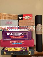 Stack of colorful board game boxes including Guess Who, Balderdash, Pictionary Junior, Frustration, Backgammon, Cribbage games and a cardboard box labeled Activision Trivial Pursuit cards, plus a black cylindrical container labeled Magnetic Dart Board with darts.