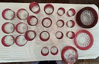 Overhead view of all 28 pieces arranged and showing red stained rims and clear thumbprint pattern on glassware with plates stacked on the side.