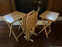 Four light-colored wooden foldable tray tables arranged around a wooden storage base on a hardwood floor in front of a fireplace.