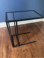 Side angled view of the black metal framed C-shaped side table with clear glass top on wood floor and blue wall background.
