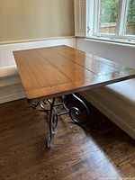 View of the dining table showing the solid maple wood top and wrought iron scrollwork base under natural lighting by a window.