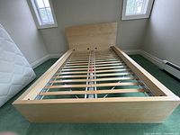 Full view of IKEA double bed frame with headboard and wooden slats in light wood finish in a room with green carpet.