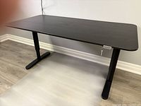 Black electric standing desk fully assembled in a room with wood laminate floor and white wall.