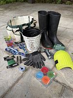 All items arranged on stone surface: tote bag, boots, gloves, knee pads, flower bucket, hand tools, hose sprayer, candles, and wires