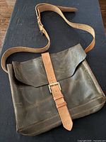 Front view of dark brown leather soft-sided satchel with tan leather strap and brass buckle closure on black background.