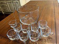 Full set of clear glass punch bowl, 8 matching cups with handles, and a glass ladle displayed on wooden table.